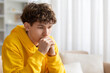 © Anastasiya - Thoughtful teenage boy sitting on sofa with hands near face, wearing yellow hoodie at home, deep in thought, worry, reflection, and quiet emotional moment indoors
