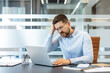 © Liubomir - Stressed businessman in glasses holding his head, experiencing a headache and frustration while working on a laptop at his office desk, dealing with professional burnout and a challenging problem