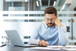 © Liubomir - Stressed businessman experiencing frustration and fatigue while reviewing paperwork and calculating data, sitting at a modern office desk with a laptop