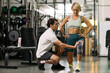 © DragonImages - Caucasian middle aged woman standing while male physical therapist kneeling and examining knee with support brace during rehabilitation session in gym setting