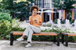 © deagreez - African American woman relaxing outdoors in city center, talking on her phone with coffee on a bench in sunny urban daylight