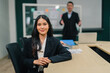 © Exnoi - Young Asian businesswoman smiles confidently, sitting at a boardroom table with a presenter blurred in the background, professionalism, leadership potential, and corporate success