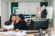 © Exnoi - Two Asian businesswomen lean close at a shared desk, one whispering a secret while reviewing documents, confidential communication, rumors, and workplace human resources issues