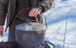 © T.Den_Team - Person preparing hot food outdoors during winter using a metal cauldron over open fire. Close view of hands cooking a meal in cold weather