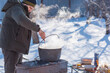 © T.Den_Team - Person preparing hot food outdoors during winter using a metal cauldron over open fire. Close view of hands cooking a meal in cold weather