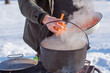 © T.Den_Team - Person preparing hot food outdoors during winter using a metal cauldron over open fire. Close view of hands cooking a meal in cold weather