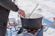 © T.Den_Team - Person preparing hot food outdoors during winter using a metal cauldron over open fire. Close view of hands cooking a meal in cold weather