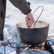 © T.Den_Team - Person preparing hot food outdoors during winter using a metal cauldron over open fire. Close view of hands cooking a meal in cold weather