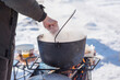 © T.Den_Team - Person preparing hot food outdoors during winter using a metal cauldron over open fire. Close view of hands cooking a meal in cold weather