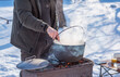 © T.Den_Team - Person preparing hot food outdoors during winter using a metal cauldron over open fire. Close view of hands cooking a meal in cold weather