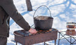 © T.Den_Team - Person preparing hot food outdoors during winter using a metal cauldron over open fire. Close view of hands cooking a meal in cold weather