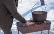 © T.Den_Team - Person preparing hot food outdoors during winter using a metal cauldron over open fire. Close view of hands cooking a meal in cold weather