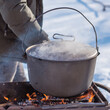 © T.Den_Team - Person preparing hot food outdoors during winter using a metal cauldron over open fire. Close view of hands cooking a meal in cold weather