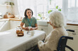 © shurkin_son - Selective focus of caregiver therapist counseling her old female disabled patient with alzheimer in wheelchair both sitting at kitchen table, senior lady pictured with back turned at camera