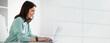 © Prostock-studio - A woman sits at a table using a laptop. She is focused on her work and smiling. The room has a simple design with light colors and natural light coming in.