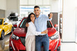 © Prostock-studio - Beautiful loving muslim family young arab man and long-haired woman buying new car in salon, standing over red luxury automobile, hugging and showing key from their new auto, copy space