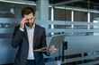 © Liubomir - Stressed businessman in a suit suffering from a headache and work pressure while holding a laptop, standing by a frosted glass partition in a modern office environment
