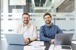 © Liubomir - Two male colleagues smiling at camera while collaborating over laptops and documents in a bright modern office, conveying teamwork, partnership and professional confidence