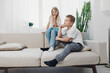 © SHOTPRIME STUDIO - Two children sit on a beige couch in a bright living room, showing thoughtful and pensive expressions. Concept of sibling emotions and family dynamics.