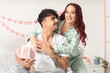 © Pixel-Shot - Young romantic couple exchanging gift box at home, smiling woman hugging man in cozy bedroom with heart decorations, valentine's day celebration, love, relationship and surprise present