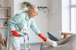 © Pixel-Shot - Mature woman with walker and pp-duster cleaning armchair at home
