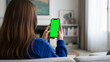 © MD Rashedul Islam - Hand holding mobile smartphone mockup with blank green screen for chroma key, rear view of woman in blue sweater using device with two hands at cozy home living room with bookshelf