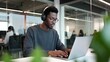 © Gennyson Marinho - A focused young Black man wearing headphones and glasses works intently on a laptop in a modern office.
