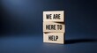 © komal - Wooden blocks stacked vertically with the reassuring message we are here to help printed clearly in black bold font against a dark blue background symbolizing support and customer service commitment