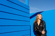 © VISTA by Westend61 - Cheerful woman in leather jacket smiling by vibrant blue wall outdoors