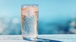 © Formoney - A clear glass filled with sparkling water sits on a wooden table. The bright blue ocean sparkles in the background on a sunny day, creating a refreshing scene.