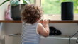 © Marco - Toddler stands on radiator beneath window, hands gripping wooden ledge, looking outside with plants and smart speaker nearby in cozy indoor space