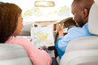 © Prostock-studio - Enjoying Travel Concept. Back view of smiling African American couple holding and looking at paper map sitting inside luxury car, choosing best way to their trip destination, sun flare