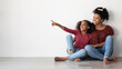 © Prostock-studio - Cheerful african american young woman mother and school girl daughter in similar outfits sitting on floor at home, hugging, smiling, teen girl pointing at copy space for advertisement, panorama