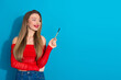 © deagreez - Beautiful young woman holding a pen on a blue background, smiling while wearing a red stylish top and denim jeans