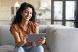 © Prostock-studio - Portrait of smiling young woman using tablet, sitting on couch at home, copy space. Happy millennial female looking at touch pad screen, browsing internet, watching video, indoors