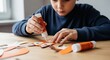 © Вадим Перегудов - Young boy carefully gluing paper cutouts together on a wooden table, focusing on a craft project. This activity promotes creativity, fine motor skills, and engaging in a childhood hobby.