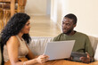© Wavebreak Media - Diverse couple sitting on beige sofa at home, using laptop and tablet, wearing casual clothes