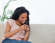 © wavebreak3 - African American woman sitting on beige sofa in living room holding black phone in pink blouse