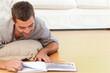 © wavebreak3 - Man lying prone on wood floor in living room, propping on beige cushion, turning magazine pages