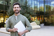 © Liubomir - Smiling man holding a portable computer and looking at camera, standing in a contemporary urban setting with a modern glass building in the background
