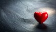 © Shahzain - Single shiny red heart-shaped object placed slightly off-center on textured dark gray surface, illuminated from upper right with soft shadow.