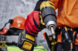 © vadish - Construction worker wearing protective red work gloves and orange high-visibility overalls holds a yellow cordless electric drill. Professional power tools are strapped to his belt.