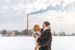 © shymar27 - Young woman in a fur coat holds a small dog in her arms, snowy landscape with a factory chimney emitting smoke in the background