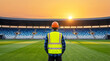 © Love 4 YOU - A lone construction worker wearing a hard hat and reflective vest stands on the field of an empty stadium during a vibrant sunset