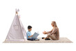 © Ljupco Smokovski - Woman sitting on a carpet with crossed legs and talking to a boy with a book in front of a kids tent