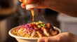 © Alodie - Hands hold a steaming bowl of noodles grilled meat and vegetables A bottle dispenses orange sauce onto the dish with a drop suspended in mid-air