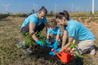 © kanpisut - Family helping planting tree in garden for save world on wind turbine field background. Eco environment concept. Family wearing blue shirts with the word volunteer planting tree on Windmill field