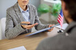 © Studio Romantic - US visa interview at consulate, officer reviewing applicant documents at office desk with United States of America flag. Female lawyer with clipboard and man discuss USA immigration application