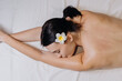 © wedmoments.stock - Close-up of a woman with dark hair and a white flower in her hair, lying on a white spa bed, enjoying a relaxing treatment in a serene spa environment