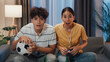 © tirachard - Asian new marriage couple sit on sofa excited cheering watching football sport game on television in living room at home. Young couple sport fans hold football watching online soccer on TV.